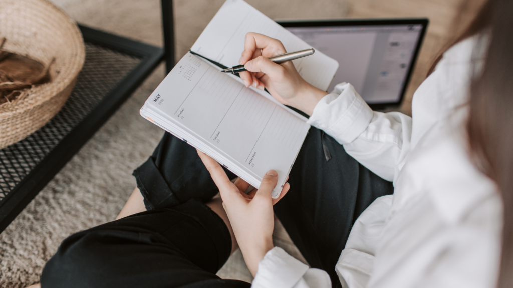 Person sitting on the floor with a notebook and pen, planning their week. The planner is open to a page for May, while a laptop rests nearby in the background, suggesting they are working on organizing or managing tasks.