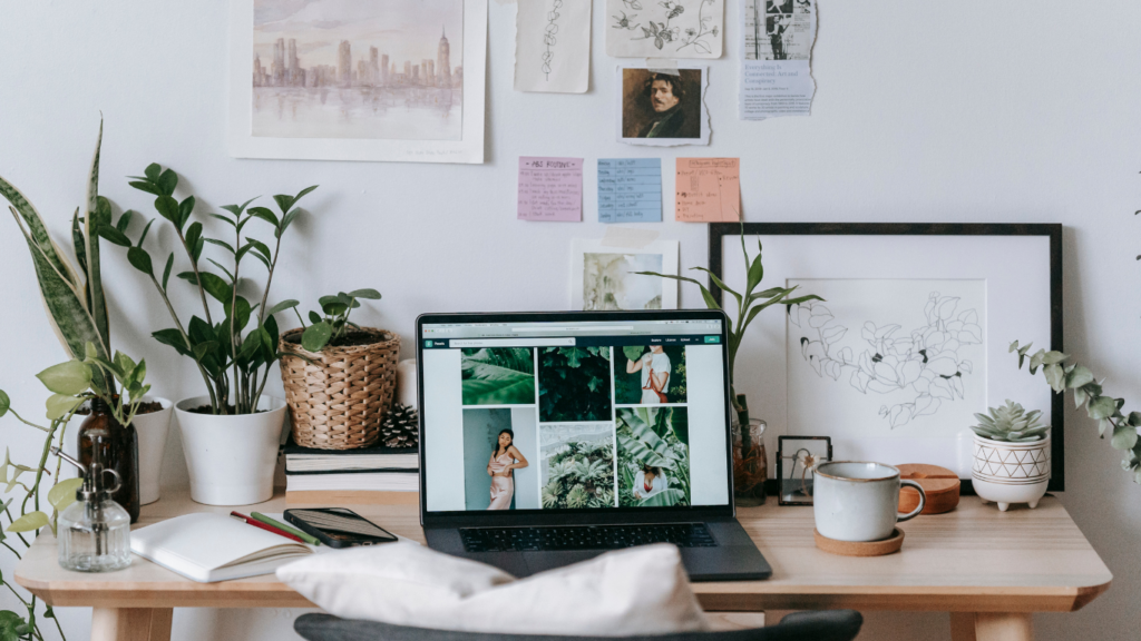 Cozy home office workspace with a laptop displaying a photo gallery on the screen. The desk is decorated with plants, a coffee mug, a notebook, and other personal items. The wall behind the desk is adorned with art prints, sketches, and colorful notes, creating a creative and calming atmosphere.