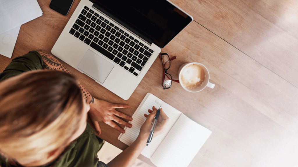 A person writing in a to-do list next to a laptop and a cup of coffee, focusing on managing daily tasks and staying organized.