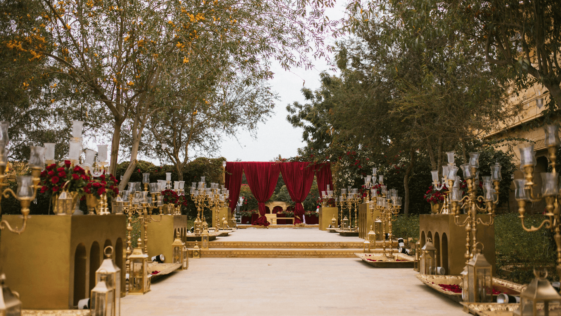 Luxury outdoor wedding ceremony with gold candelabras, ornate lanterns, red velvet draping, and elaborate cultural decorations for elegant celebrations