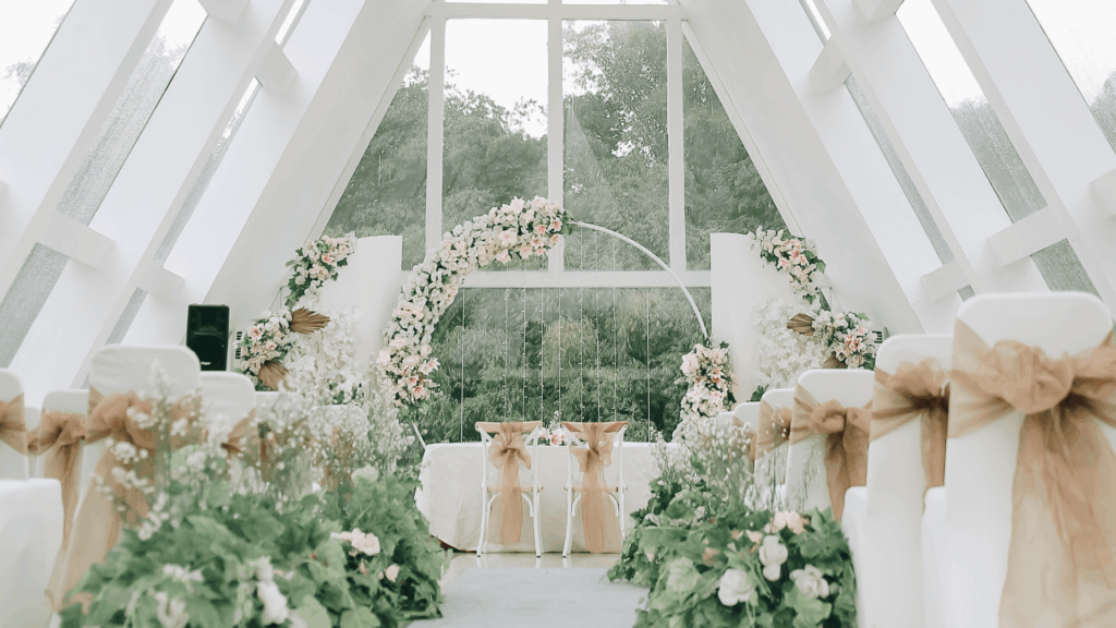 Bright modern chapel wedding venue with white architectural beams, floor-to-ceiling windows, pink and white floral arch, and romantic garden ceremony setup