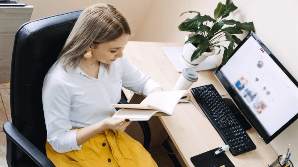 A woman in a bright yellow skirt reviewing notes at her desk. A computer screen showing social media content is visible alongside a clean and modern workspace.