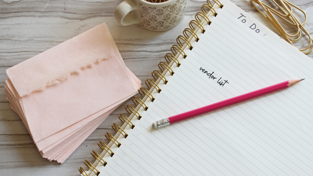 A pink notebook open to a “To Do” list, with “preferred vendor list” written in bold letters. A cup of coffee and decorative paper sit on a marble tabletop, creating a cozy planning atmosphere.
