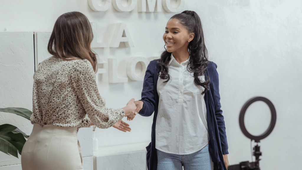 Two women shaking hands in a professional setting, smiling as they meet. The connection symbolizes forming partnerships that could lead to being included on a preferred vendor list. The background features light decor and a ring light setup, suggesting a creative business environment.