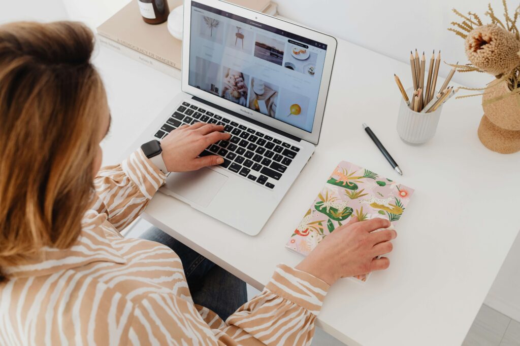 A woman working at her desk with a laptop open to a portfolio website, holding a colorful notebook. A neatly organized workspace with pencils and decor in the background.