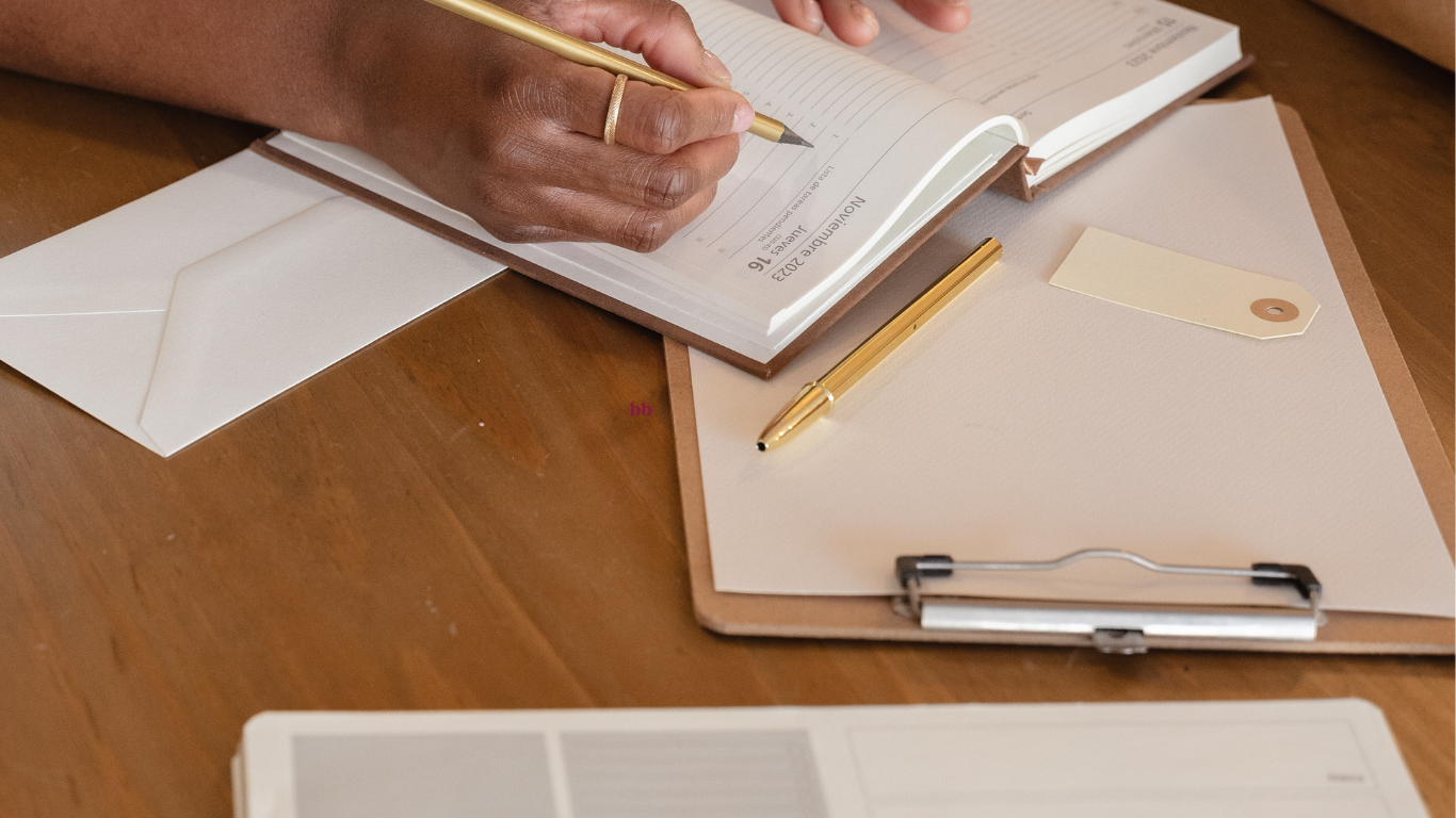 A woman of color writing in a planner at a wooden desk, surrounded by office supplies and documents. Her focus suggests she is organizing and strategizing for a project.