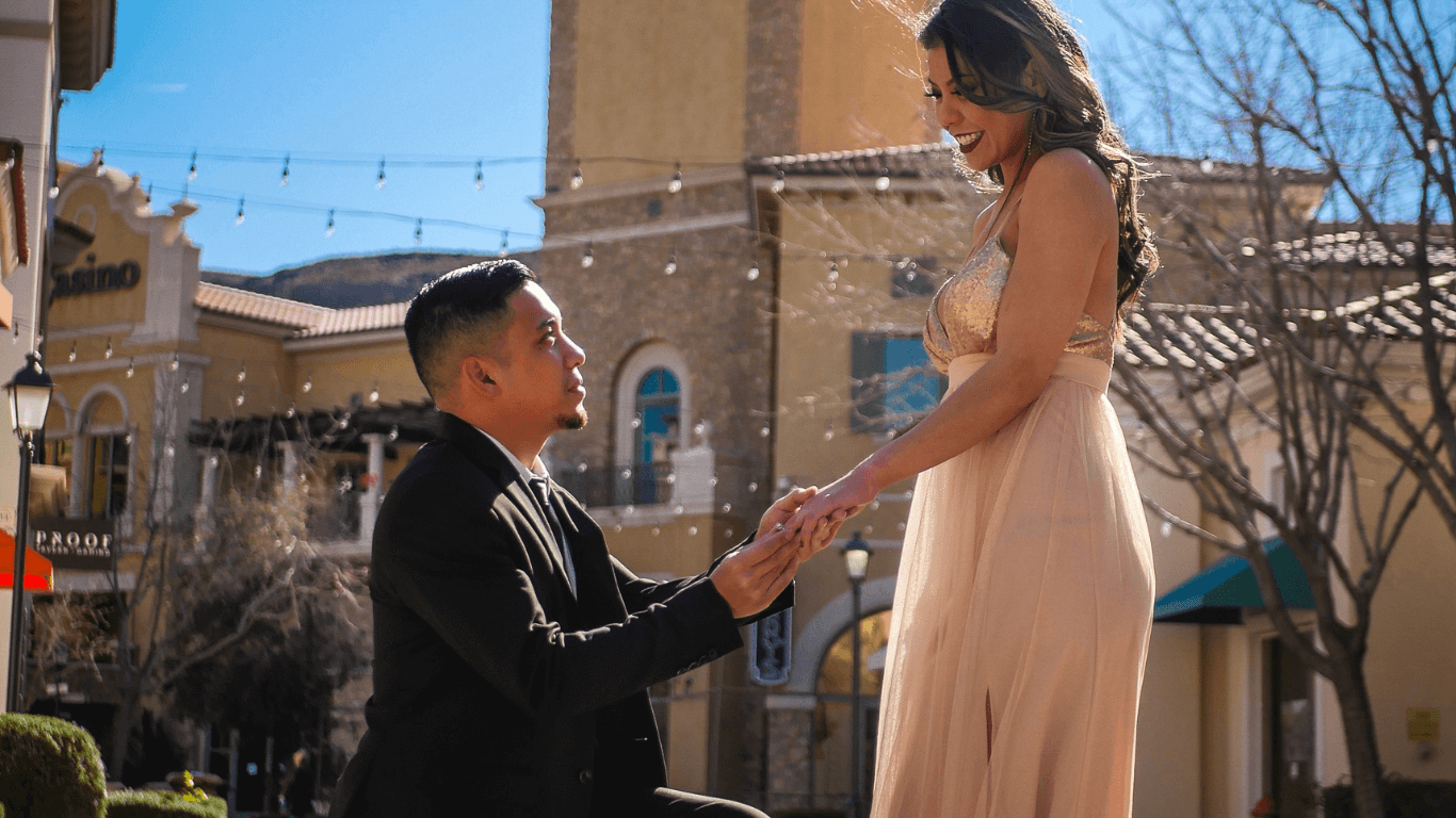 A man on one knee proposing to a woman in a beautiful outdoor setting, with sunlight casting a warm glow on the scene. The woman, dressed elegantly in a blush pink dress, smiles as she accepts the proposal.