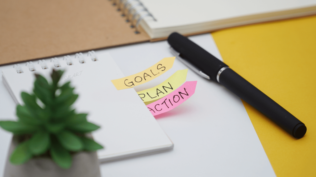 Sticky notes labeled Goals, Plan, and Action next to a notebook and pen on a desk.