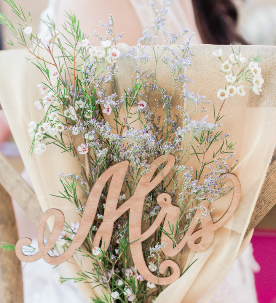 A close-up shot of a wooden "Mrs" sign attached to the back of a chair, adorned with delicate white and lavender flowers and greenery. The arrangement is beautifully complemented by a soft, sheer fabric draped over the chair, creating an elegant and romantic wedding decor.