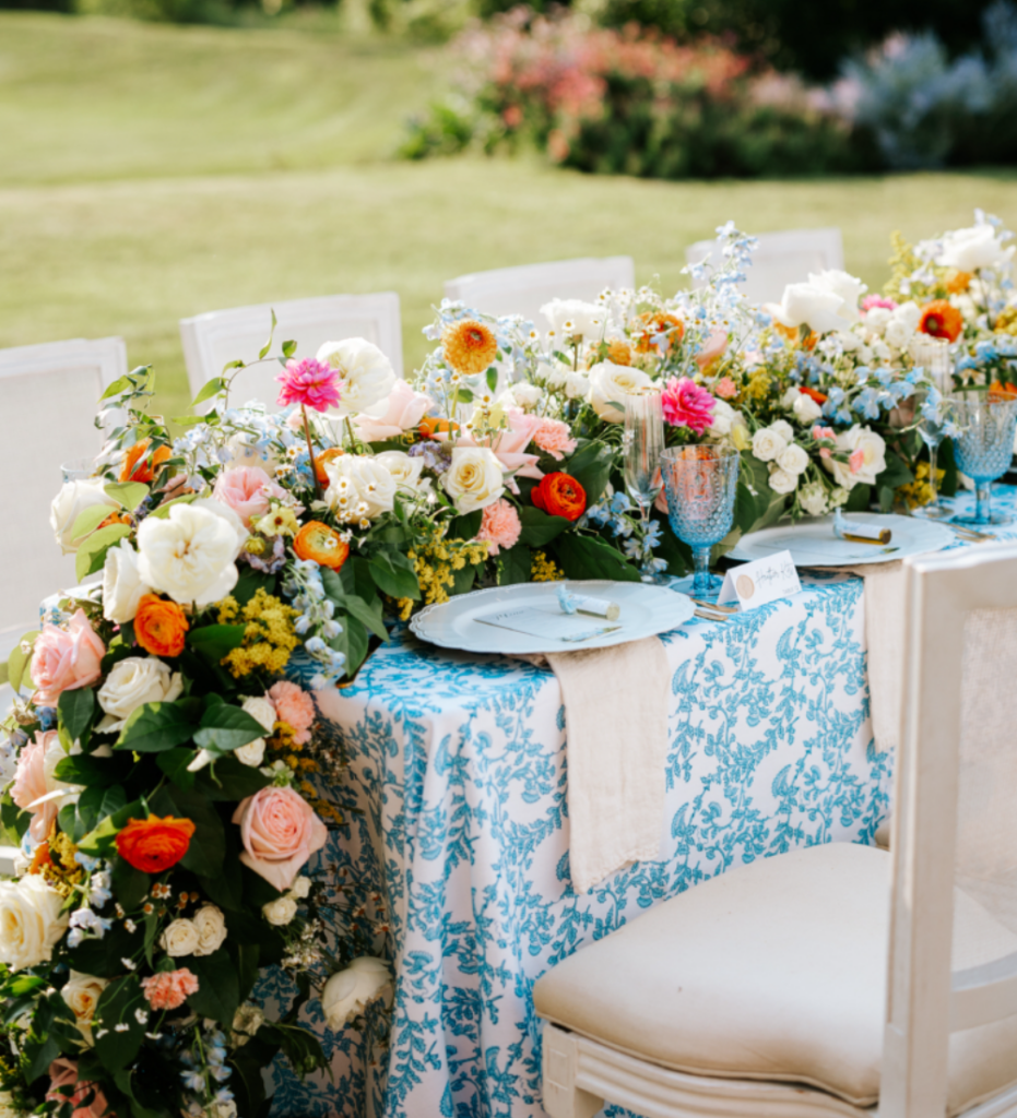 Outdoor styled shoot reception table draped in elegant blue and white linens, adorned with a cascade of vibrant flowers flowing down the table’s edge, adding a touch of romance and luxury to the setting.