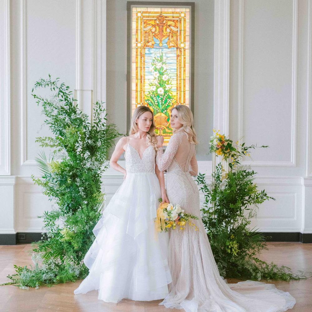 The image shows two women standing side by side in a beautifully decorated room. Both are wearing elegant bridal gowns, one in a white dress with a tiered skirt and the other in a light-colored lace gown. They are positioned in front of a stained glass window, which adds a vibrant and artistic backdrop. The space around them features lush greenery, adding a natural, serene atmosphere to the setting.