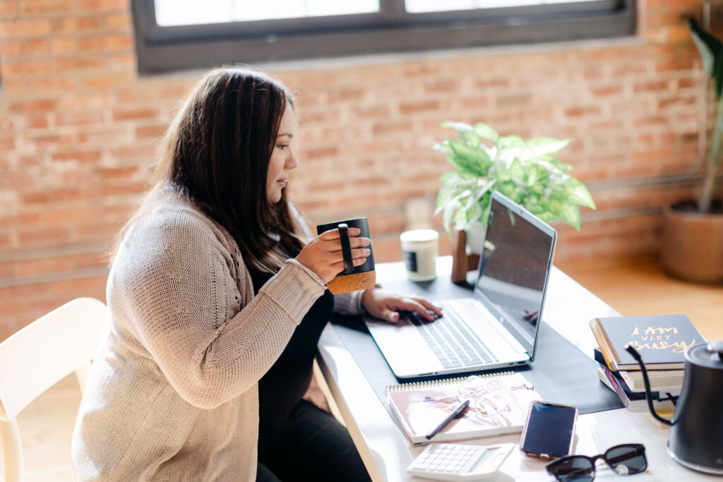 OBM drinking coffee while working on a laptop at a desk with a notebook, phone, and other office supplies.