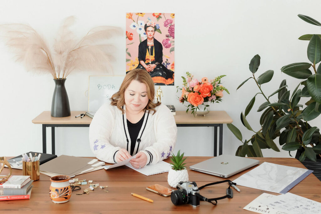 Cinthia Onines, founder of Boda Bliss and Marketing Systems Architect, working at her desk with a laptop, camera, notebooks, and stationery.