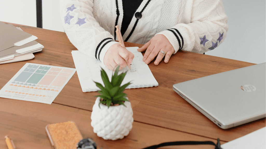 Hands writing in planner beside laptop and files, symbolizing workflow planning for creative businesses.