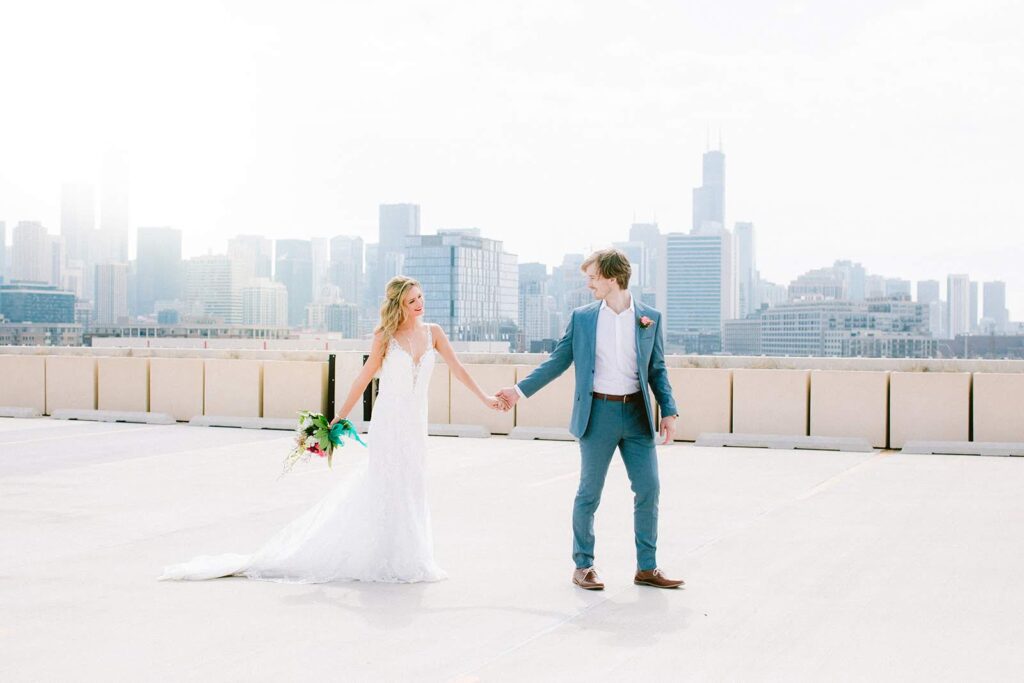 Bride and groom holding hands on a rooftop with a stunning city skyline backdrop, capturing an intimate moment on their wedding day.