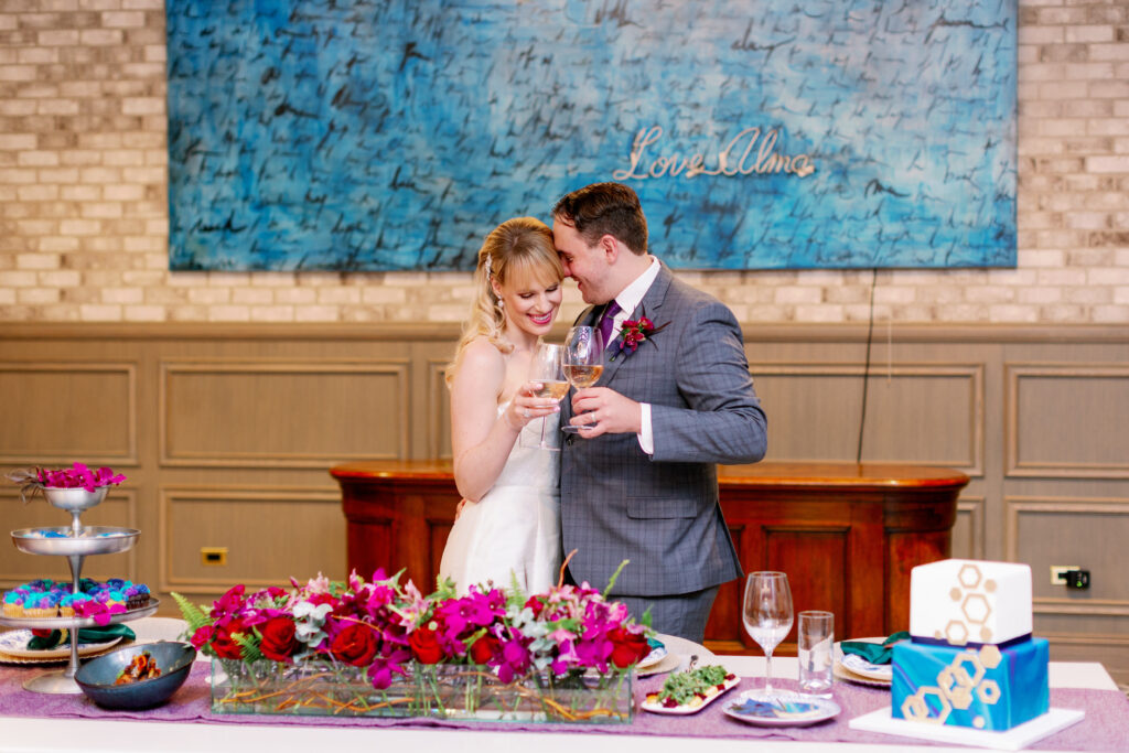 Bride and groom share a toast in a beautifully decorated reception area with a vibrant floral centerpiece, celebrating their love with elegance and joy.