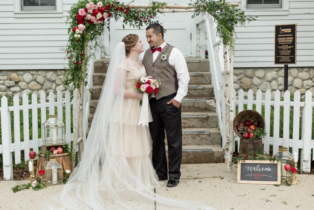 "Bride and groom standing by a charming rustic wedding setup with a wooden arch adorned with red and pink roses, against a vintage white house backdrop.