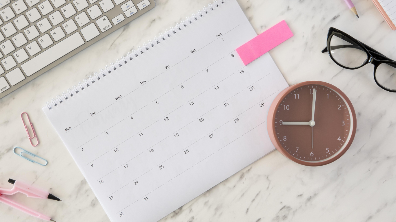 A calendar and clock on a desk with colorful office supplies, symbolizing time management and planning for achieving business goals within a 12-Week Year framework.