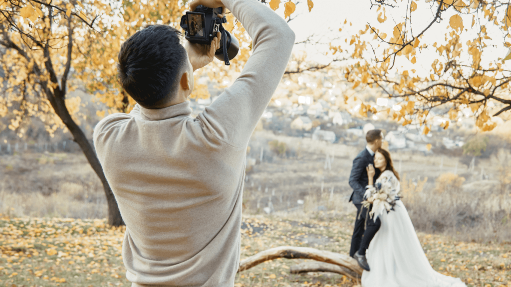 A wedding photographer taking a picture of a couple outdoors in an autumn setting, showcasing the photographer at work during a wedding shoot.