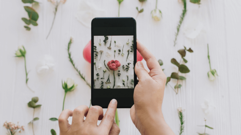 Close-up of a person using a smartphone to take a photo of floral arrangements, representing social media marketing their photography business. 