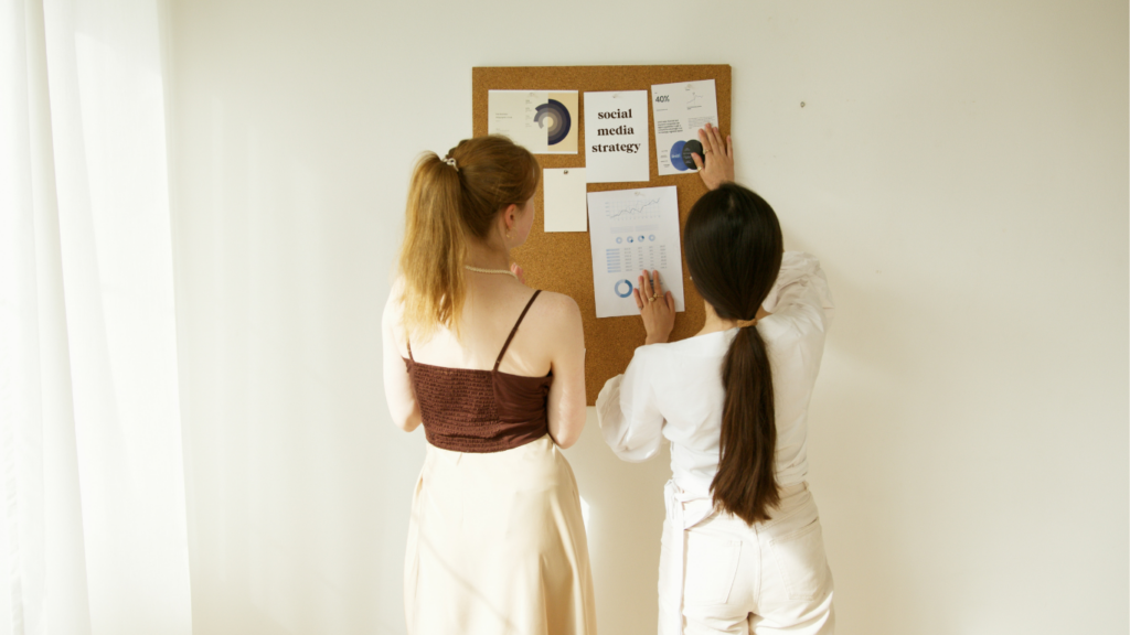 Wedding Planner and Online Business Manager collaborating on a corkboard filled with social media strategy charts and graphs, planning and organizing content visually.