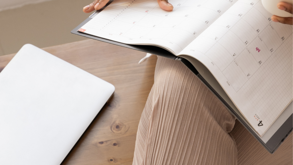 Close-up of a person holding a planner and reviewing schedules with a laptop in the background.