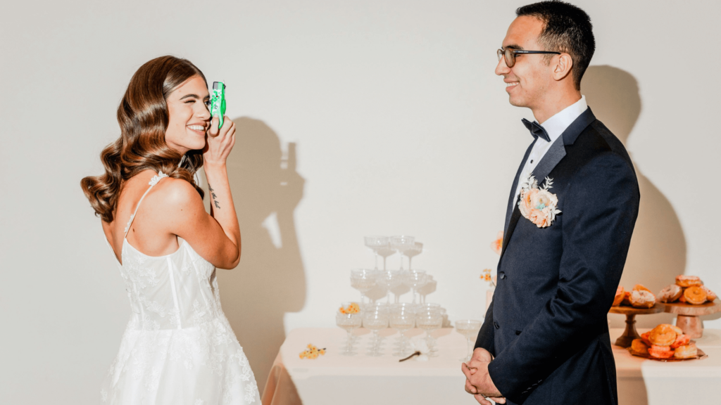 A smiling bride playfully holding a camera while the groom stands nearby at their wedding reception, surrounded by elegant décor.