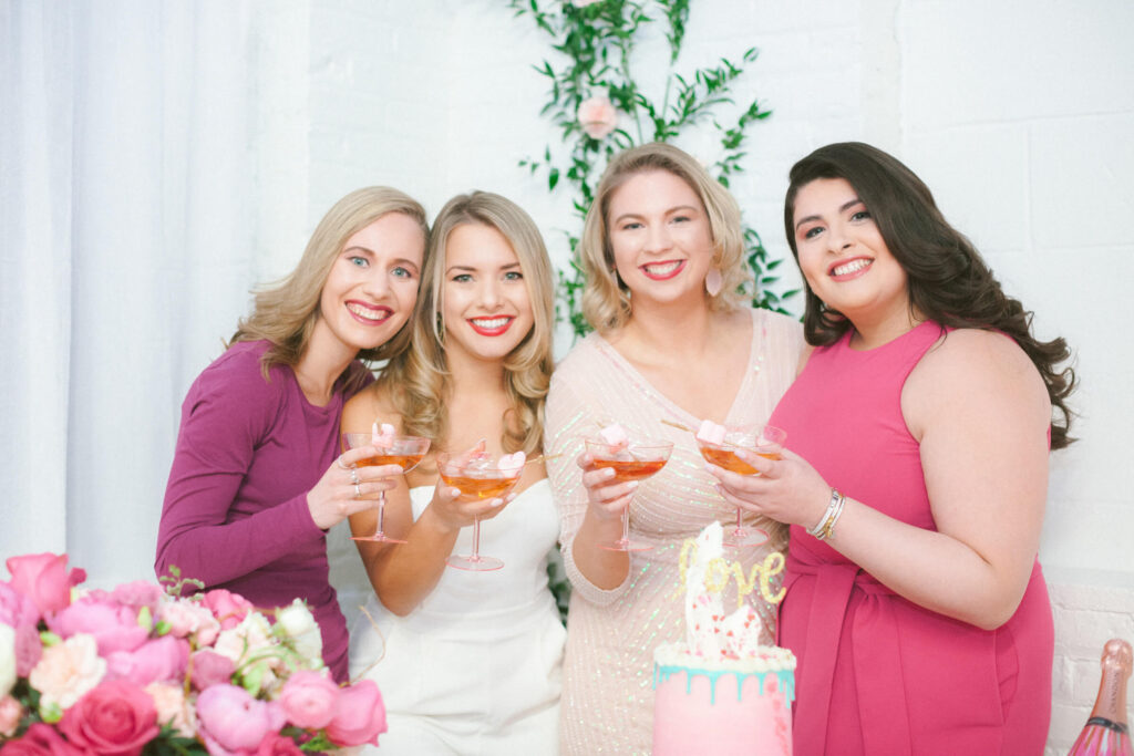 A group of four smiling women dressed in pink, white, and blush tones clinking glasses at a beautifully decorated table with a pink and gold cake, celebrating friendship and business connections at a Galentine’s Day referral networking event.