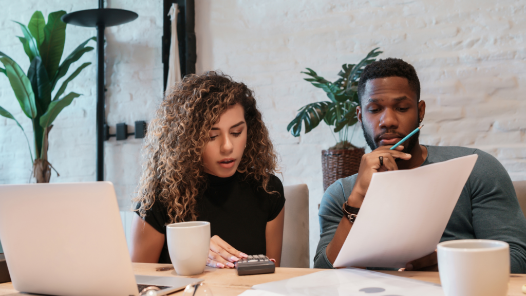  A couple reviewing wedding planning documents with a laptop and calculator, making decisions about vendor listings on platforms like Zola and The Knot.