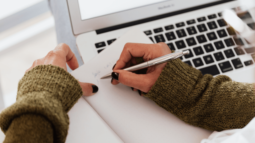 A wedding professional writing notes in a dotted planner while using a laptop to refine her CRM workflow.