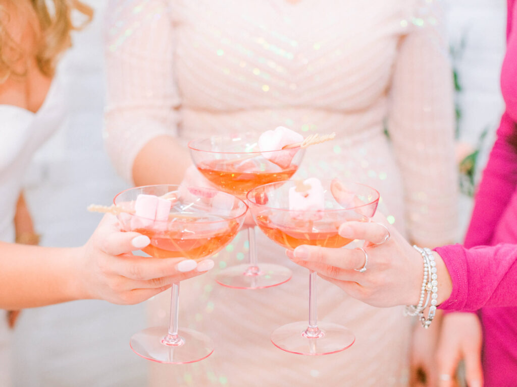 Close-up of three elegant cocktail glasses filled with a sparkling pink drink, garnished with pastel pink marshmallows on skewers, as a group of women toasts together in celebration of Galentine’s Day.