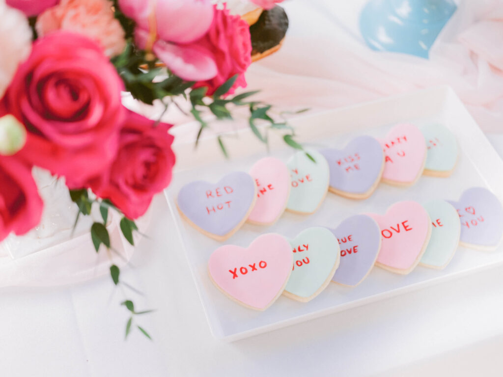 A tray of heart-shaped pastel cookies decorated with sweet messages like 'XOXO,' 'Kiss Me,' and 'Red Hot,' placed next to a vibrant pink and white floral arrangement, perfect for a Galentine’s Day networking event.