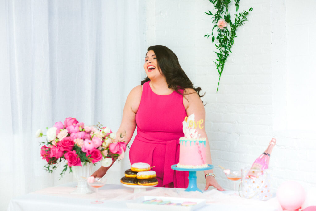 A woman in a bright pink dress laughing joyfully at a beautifully decorated dessert table with pink and gold accents, featuring a cake with 'love' topper, fresh flowers, and an array of sweet treats for a festive Galentine’s Day gathering.