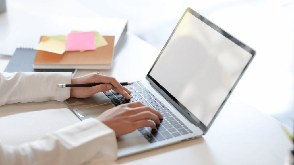 Wedding vendor typing on a laptop while managing online listings for Zola or The Knot with notebooks and post-its nearby.