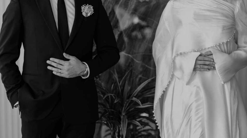 Black and white close-up of bride and groom's hands showing wedding rings, with bride's detailed beaded dress visible.
