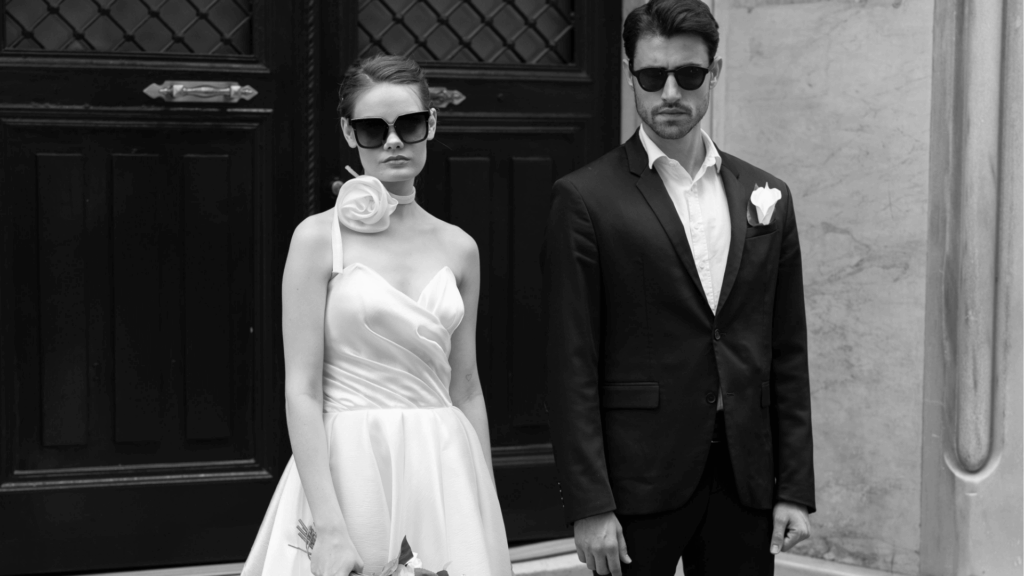 Black and white photo of a stylish couple wearing sunglasses against a dark architectural background, bride in white wrap dress and groom in black suit.
