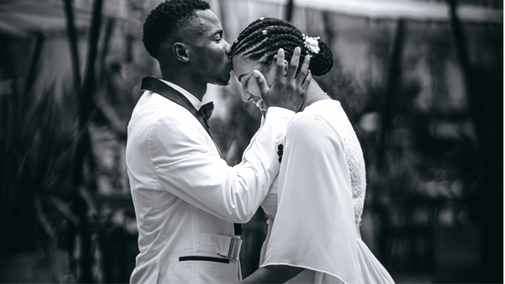 Black and white close-up of a groom kissing the bride's forehead while she smiles, with detailed white wedding dress visible.