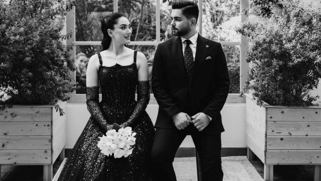 Black and white photo of a couple in modern formal wear, bride in a black beaded gown and groom in a dark suit, holding a white bouquet in a modern venue.