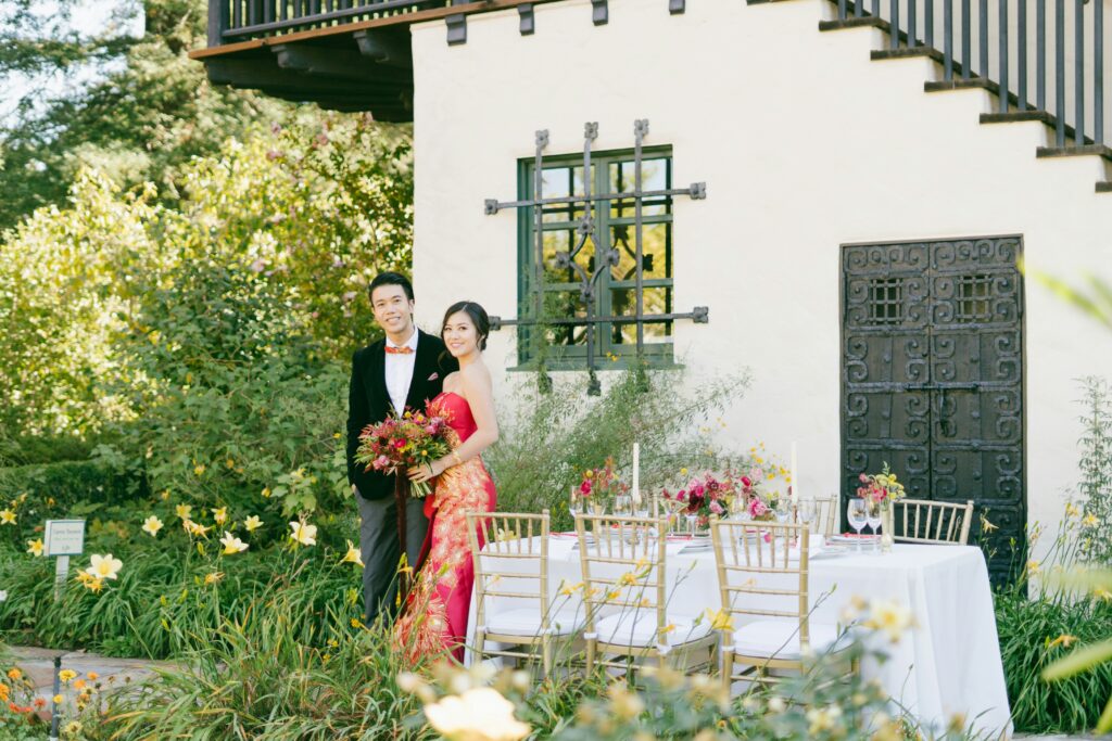 Asian wedding couple in elegant red and black attire posing beside a garden reception table, symbolizing inclusive marketing and cultural representation in wedding storytelling.