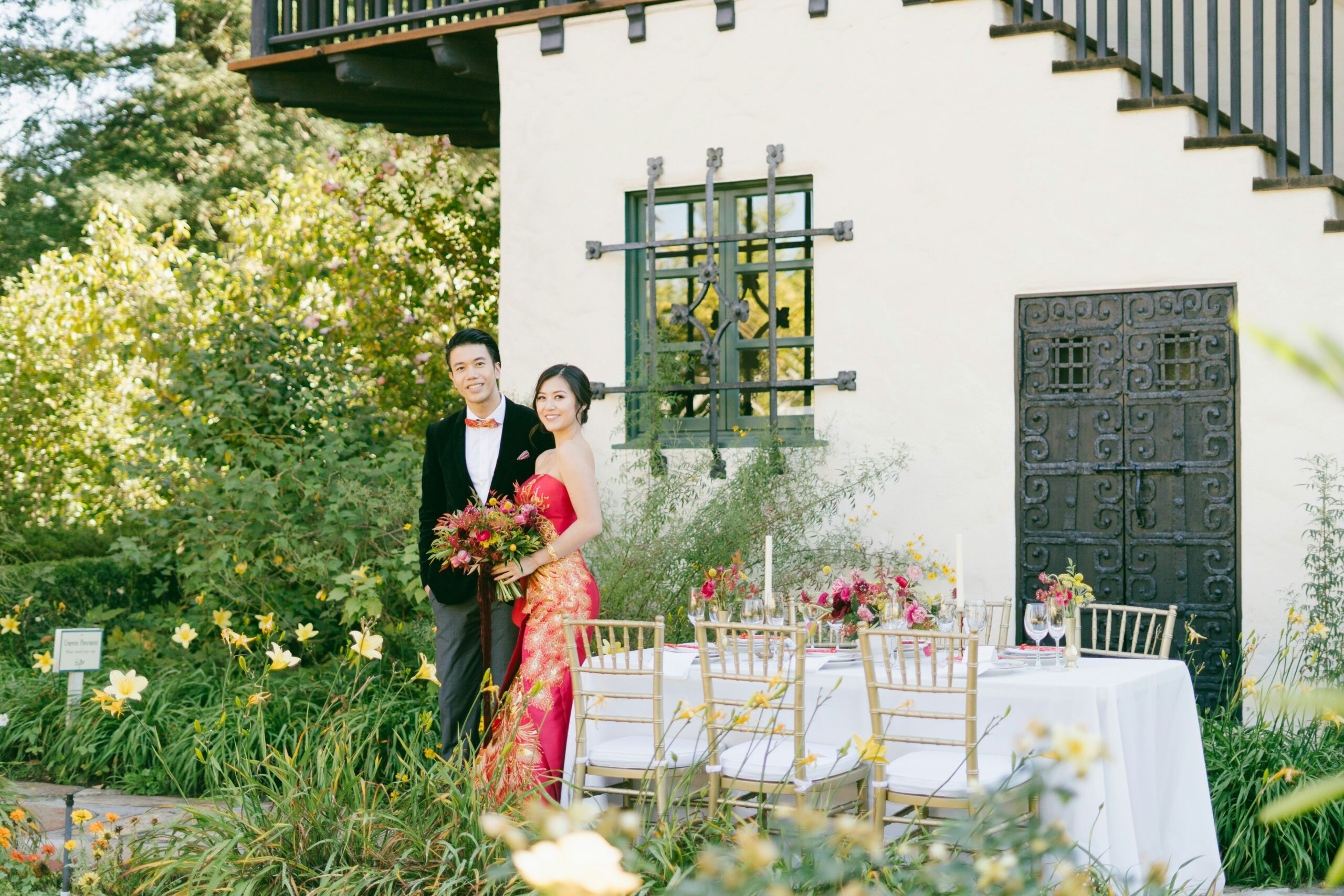 Asian wedding couple in elegant red and black attire posing beside a garden reception table, symbolizing inclusive marketing and cultural representation in wedding storytelling.