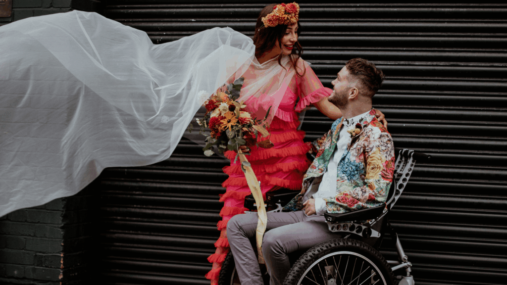 A bride in a pink ruffled dress smiling at her groom in a wheelchair, representing accessibility and disability inclusion in wedding marketing.