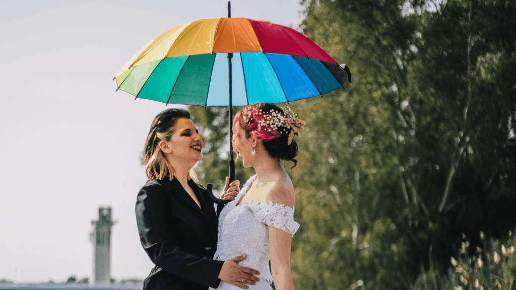 Two brides under a rainbow umbrella smiling together, representing LGBTQ+ inclusion and authentic wedding storytelling.