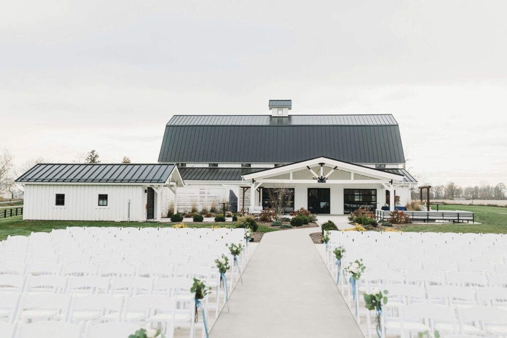 Modern white farmhouse wedding venue with black metal roof, outdoor ceremony setup, white chairs, and greenery-lined aisle