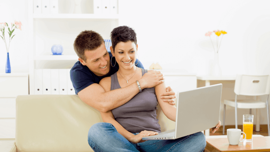 Couple sitting on a couch researching wedding vendors together on a laptop at home.